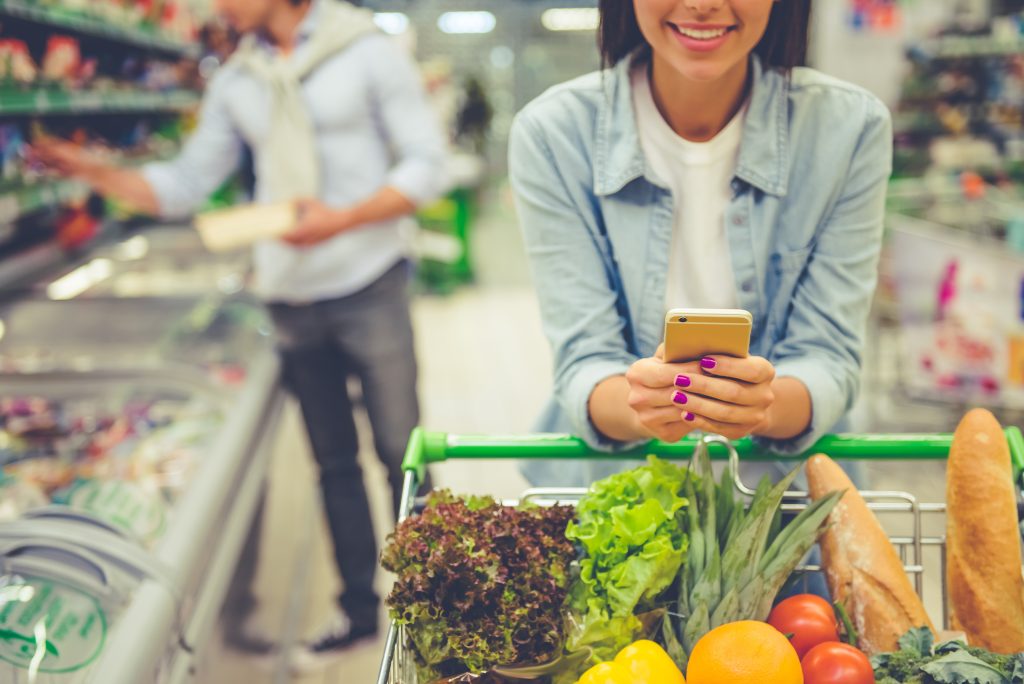 Pareja joven disfrutando de sus compras en Nueva Valdeluz (Guadalajara): mujer sonriendo con su móvil mientras su pareja elige productos en el supermercado.