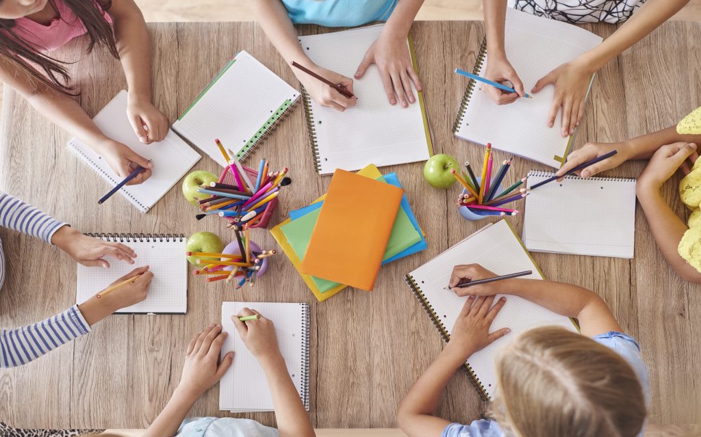 Vista cenital de un grupo de niños sentados a una mesa de madera escribiendo en cuadernos y realizando tareas escolares en Nueva Valdeluz